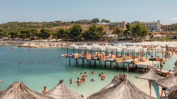 Słoneczny dzień na plaży Ksamil. Ludzie kąpią się w turkusowej wodzie, odpoczywają pod białymi parasolami na pomostach i na piaszczystym brzegu.