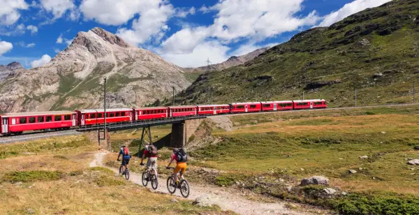Treno rosso attraversa un ponte su un paesaggio alpino mozzafiato. Tre ciclisti pedalano su un sentiero sottostante, godendosi la vista della ferrovia del Bernina.