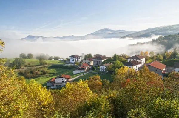 Pueblo en el **valle del Baztan** cubierto por niebla matutina, con casas blancas y tejados rojos entre árboles otoñales.