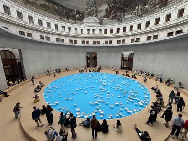 A circular room with a frescoed ceiling, featuring a bright blue pool dotted with white ceramic bowls. Visitors observe this installation, part of the François-Henri Pinault art collection.