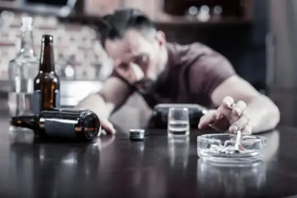 Mężczyzna z papierosem w popielniczce, butelki i kieliszek na stole. Nadmierne spożycie alkoholu może pogorszyć hemoroidy.