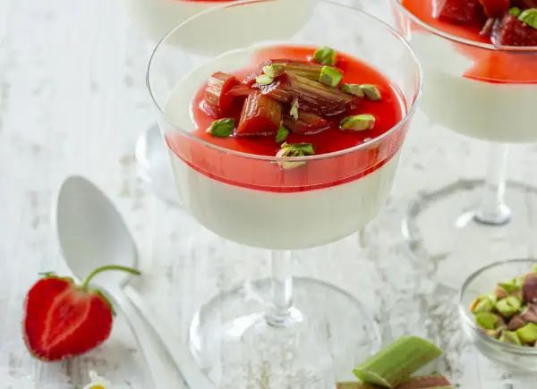 Panna cotta z rabarbarem i pistacjami w kieliszku. Słodki deser z rabarbarem, ozdobiony zielonymi orzeszkami.