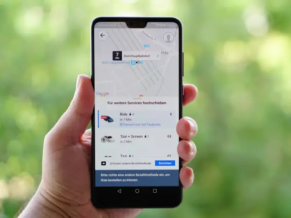 Hand hält Smartphone mit der besten Taxi App Deutschlands. Anzeige zeigt Fahrtoptionen nach Köln Hauptbahnhof.