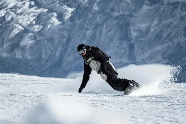 Snowboarder w kasku i goglach, z ochraniaczem nadgarstka snowboard, pochyla się nisko, tworząc obłok śniegu podczas zjazdu.