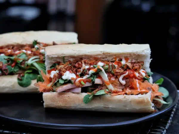 Zwei appetitliche Banh Mi Sandwiches mit Fleisch, Gemüse, knusprigen Zwiebeln und Saucen auf einem Teller.
