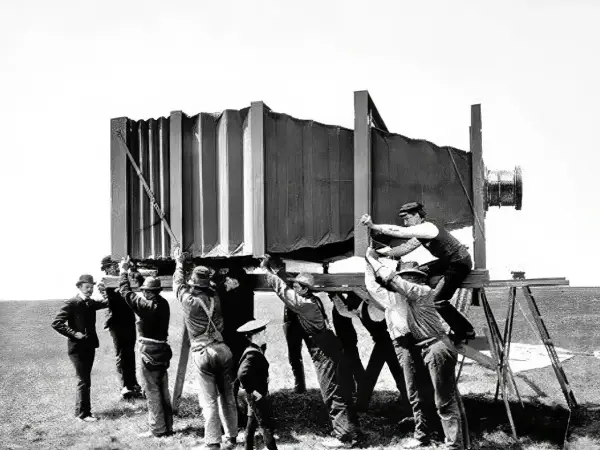Grupa mężczyzn w historycznych strojach ustawia gigantyczny aparat fotograficzny. Wyobraź sobie, kiedy wynaleziono aparat fotograficzny i jak wielkie musiały być pierwsze urządzenia!