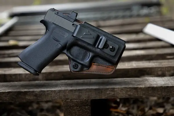 Pistolet w kaburze udowej z czarnego polimeru i brązowej skóry, z widocznym celownikiem optycznym.