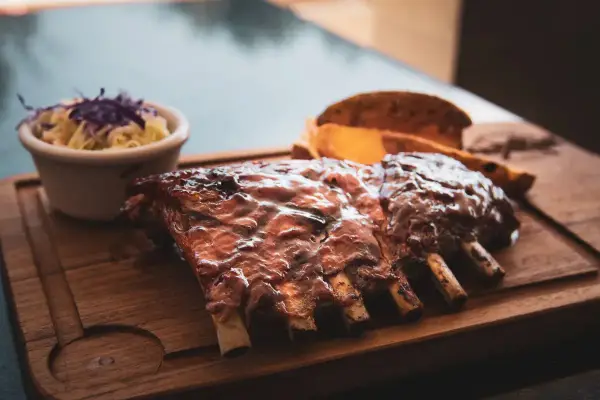 Un plat de ribs savoureux, nappés d'une sauce brillante, accompagnés de coleslaw et de quartiers de pommes de terre sur une planche en bois.