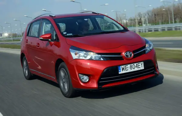 Czerwona Toyota Corolla Verso jedzie po drodze. Kierowca w okularach przeciwsłonecznych prowadzi auto.