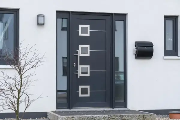 Nowoczesne drzwi zewnętrzne w ciemnoszarym kolorze z geometrycznymi przeszkleniami i metalowymi elementami. Obok skrzynka na listy.