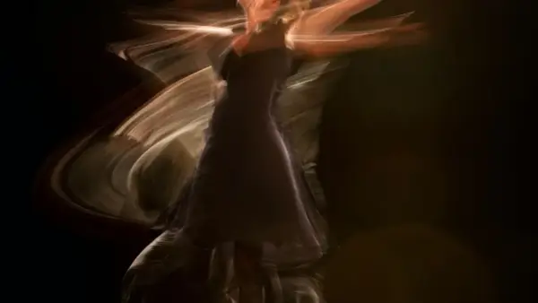 Una figura etérea en un vestido oscuro se mueve con gracia, sus brazos extendidos crean estelas de luz. Es como soñar con bailar, un instante capturado en movimiento.
