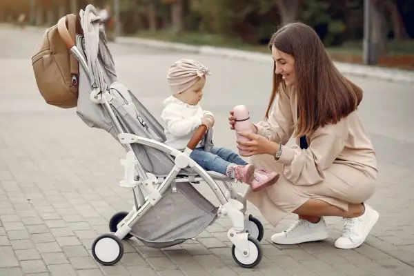 Mama karmi córeczkę z butelki, siedząc obok wózka. Idealne wózki dla dziewczynki na spacer.
