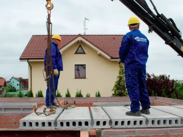 Dwóch budowlańców w żółtych kaskach i niebieskich kombinezonach montuje prefabrykowany strop. Jeden z nich stoi na płycie, drugi czeka na dalsze instrukcje.