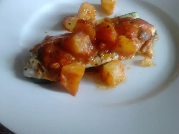 Un delicioso jargo pescado cubierto con salsa de tomate y trozos de patata, servido en un plato blanco.