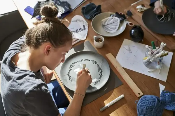 Dziewczyna maluje na talerzu, tworząc wzór. Wokół materiały do rękodzieła, jak do zimna porcelana przepis.