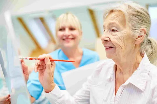 Anziana dipinge con un pennello, assistita da un'infermiera sorridente. Un esempio di terapie non farmacologiche per l'Alzheimer che stimolano la creatività.