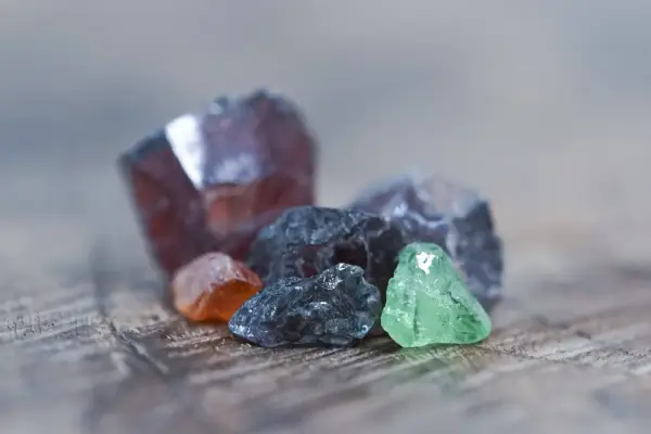 Raw gemstones in various colors, including deep red, orange, grey, and green. The deep red stone might be the birthstone for January.