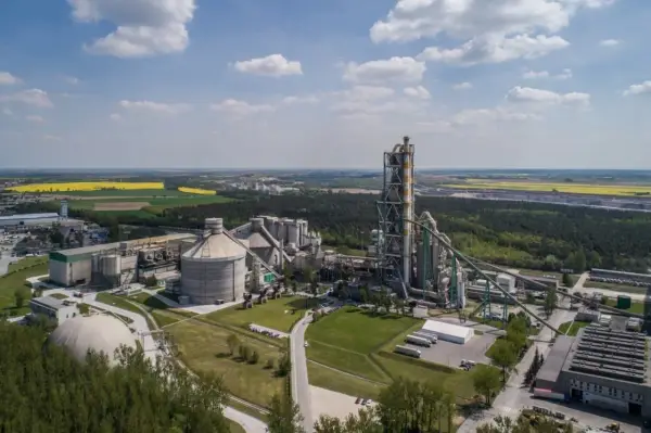 Zakłady Lafarge z lotu ptaka, z dominującą wieżą pieca cementowego i silosami, otoczone zielenią i polami.