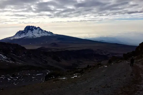 Vue sommet Kilimandjaro, paysage désertique et enneigé sous un ciel nuageux. Un randonneur solitaire sur le sentier.