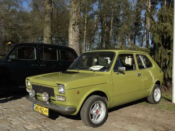 Zielony fiat 127 z lat 70. stoi obok czarnej taksówki na tle drzew.