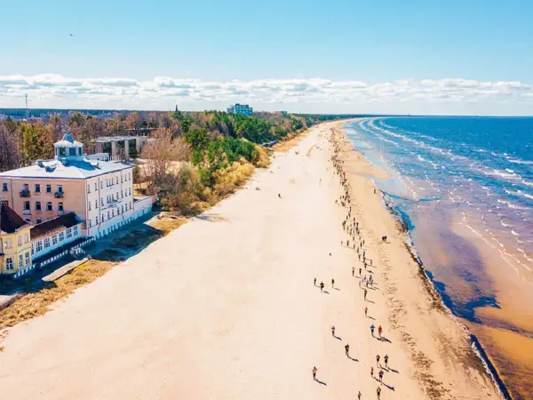 Szeroka, piaszczysta plaża na Łotwie, z budynkami przy brzegu i ludźmi spacerującymi wzdłuż morza.