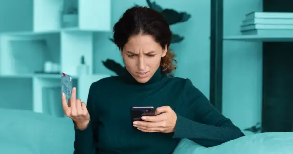 Zmarszczona kobieta trzyma kartę i telefon, zastanawiając się nad bezpieczne zakupy w internecie.