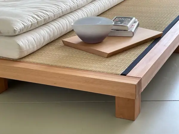 Eine gestapelte, weiße Matratze eines japanischen Bettes ruht auf einer Tatami-Matte. Daneben liegen ein Buch und eine Schale auf einem Holzbrett.