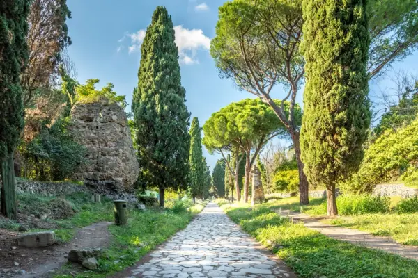 Allée pavée bordée de cyprès et de pins sur la Via Appia, avec des ruines antiques sur le côté.