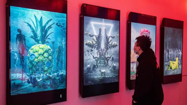 Ein Mann betrachtet digitale Kunstwerke, die an einer Wand hängen. Die Werke zeigen surreale Szenen, wie eine Ananas unter Wasser mit Skeletten und eine symmetrische Figur mit vielen Armen. Diese Werke stammen von einem berühmten Grafik Künstler.