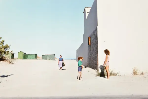 Luigi Ghirri foto: una donna cammina sulla spiaggia, due bambine giocano vicino a un edificio bianco.