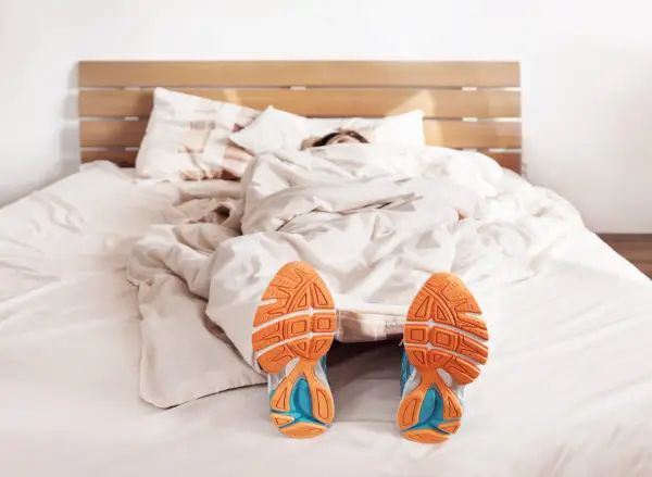 Füße mit Laufschuhen ragen aus einer Decke. Der Wecker klingelt, aber die Person träumt noch vom morgens joggen.