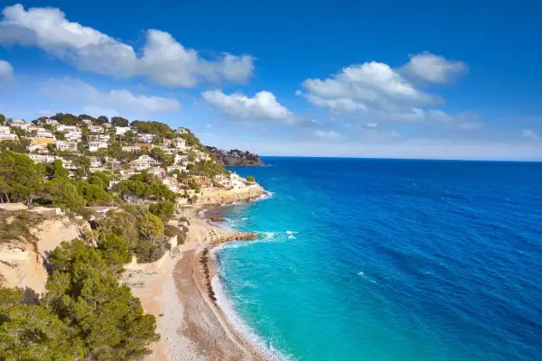 La **cala Baladrar** se asoma entre casas blancas y pinos verdes, con aguas turquesas y un mar azul intenso bajo un cielo nublado.