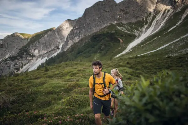 Coppia in escursioni alto adige, camminano su un sentiero verdeggiante con montagne imponenti sullo sfondo.