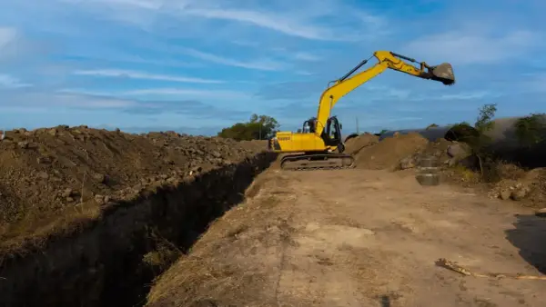 Excavadora amarilla trabajando en la creación de varios tipos de zanjas, con montones de tierra y tuberías grandes en el fondo.