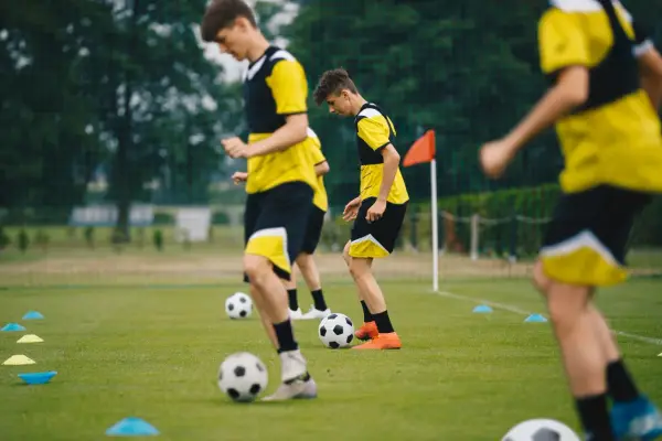 Młodzi piłkarze w żółto-czarnych strojach ćwiczą na boisku. Widać piłki i pachołki, co sugeruje trening dla różnych kategorie wiekowe w piłce nożnej.