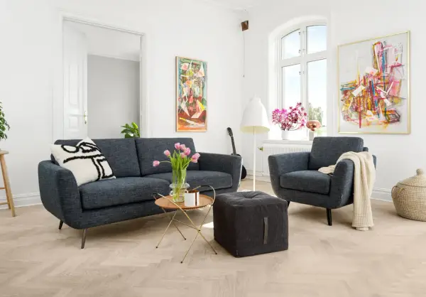 En modern soffgrupp 3+2 i mörkblått tyg, med en fåtölj, soffbord, pall och konst på väggarna.