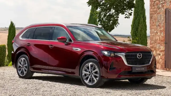Lśniąca, czerwona Mazda CX-80 zaparkowana na żwirowej drodze, otoczona zielenią i ceglanym murem.
