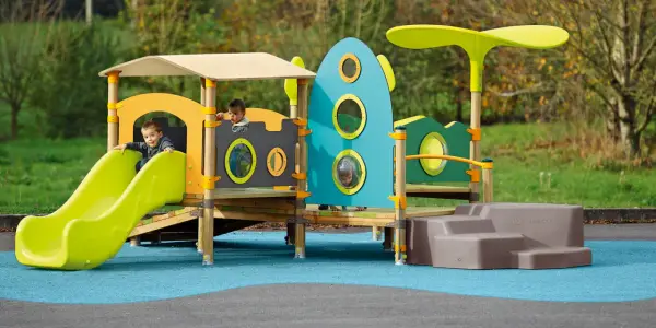 Des enfants jouent sur une aire de jeux colorée, respectant la réglementation aire de jeux extérieur. Un toboggan vert vif et une structure inspirée d'une fusée bleue.