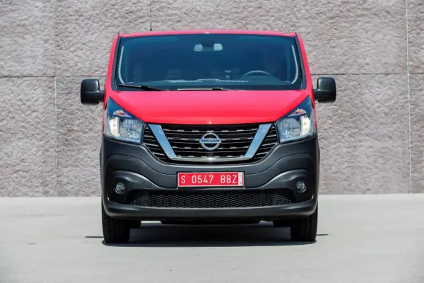 Czerwony Nissan NV300 z czarnym zderzakiem i chromowaną atrapą chłodnicy stoi przed betonową ścianą.