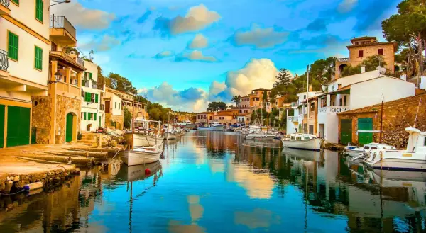 Un pintoresco pueblo pesquero con casas blancas y verdes, barcos amarrados en un canal de aguas cristalinas y un cielo azul con nubes esponjosas.