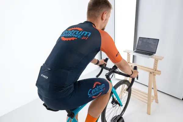 Mężczyzna w stroju kolarskim ćwiczy na rowerze stacjonarnym, korzystając z aplikacji Zwift na laptopie.