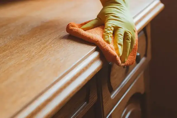 Ręka w żółtej rękawiczce czyści drewnianą komodę pomarańczową ściereczką. Dowiedz się, czym rozpuścić klej, by usunąć uporczywe plamy.
