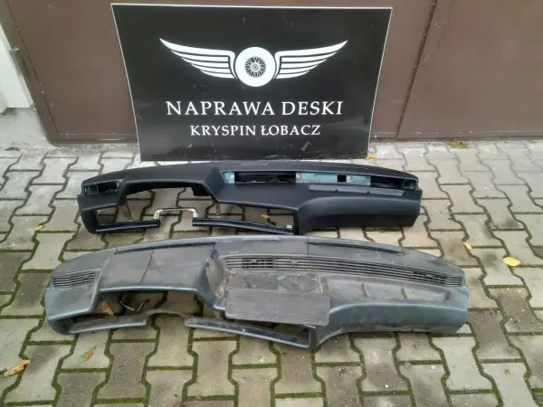 Dwa kokpity samochodowe, jeden czarny, drugi szary, leżą na chodniku przed tablicą z napisem "NAPRAWA DESKI".