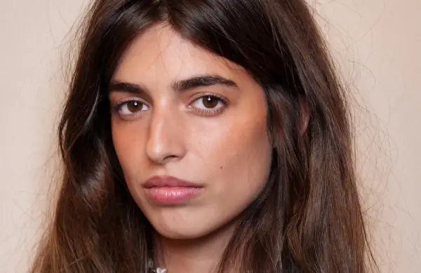 Portrait d'une jeune femme aux longs cheveux bruns. Son maquillage met en valeur ses yeux et ses lèvres, avec un soin particulier apporté à son sourcil poil à poil.