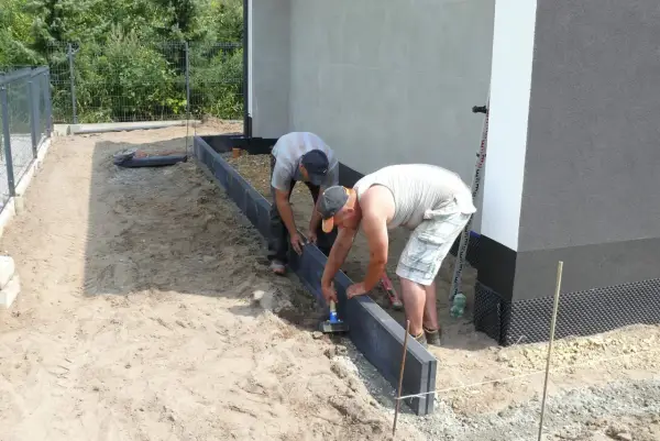 Dwóch mężczyzn układa obrzeża pod kostkę. Suchy beton pod kostkę jest przygotowywany do dalszych prac.