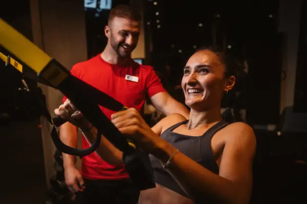Frau lacht und trainiert mit TRX-Bändern, ihr Trainer lächelt. Sie will schnell wieder fit werden.