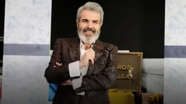 Lorenzo Caprile, con su característica barba canosa, sonríe mientras sostiene unas gafas. Viste una chaqueta de tweed con bordados florales.