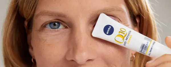 Kobieta z niebieskimi oczami, z kremem Nivea Q10 Anti-Wrinkle Power zakrywającym jedno oko. Skóra wokół oczu ma drobne kurze łapki.