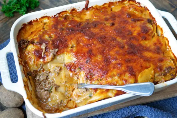 Omas Kartoffelauflauf mit Hackfleisch, goldbraun überbacken, dampft in einer weißen Auflaufform. Eine Gabel steckt in der saftigen Schicht.