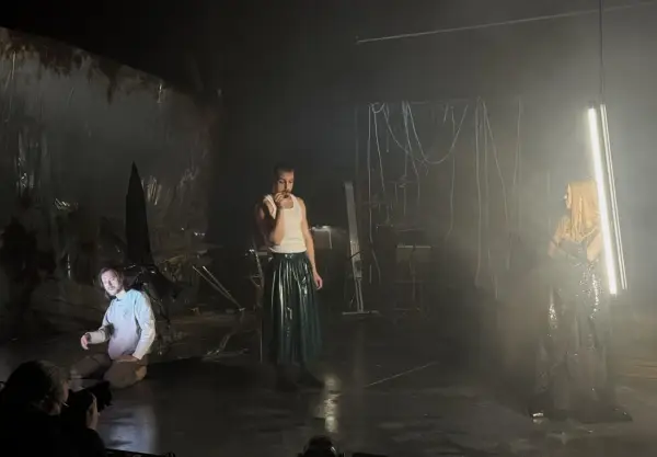 Trzej aktorzy na scenie, jeden klęczy, drugi stoi w błyszczącej spódnicy, trzecia w połyskującej sukni. Scenografia sugeruje **epic the musical**.